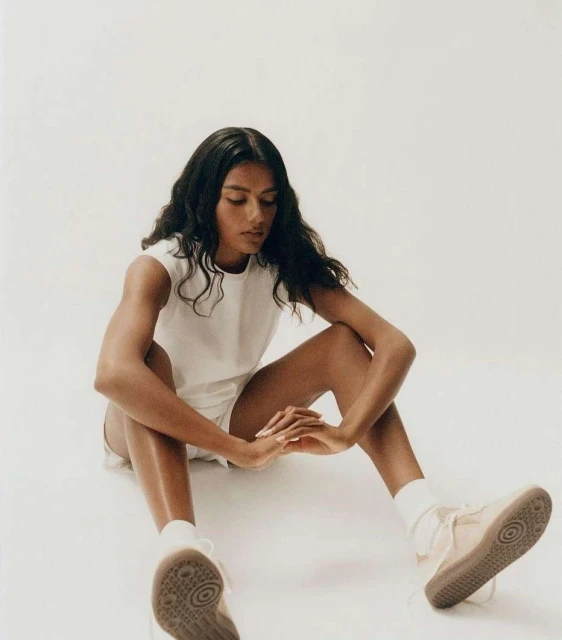 A young woman with long dark hair sits on the floor in a white shirt and shorts, wearing sneakers, in a brightly lit studio setting.
