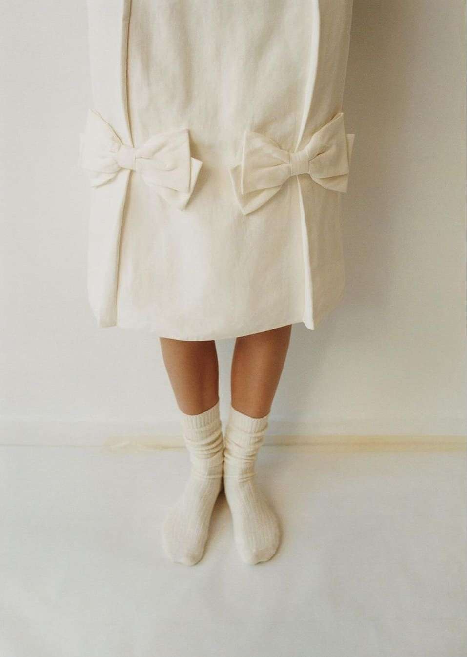 Close-up of legs in cream socks beneath a white dress with bow accents against a plain background