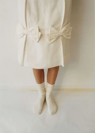 Close-up of legs in cream socks beneath a white dress with bow accents against a plain background