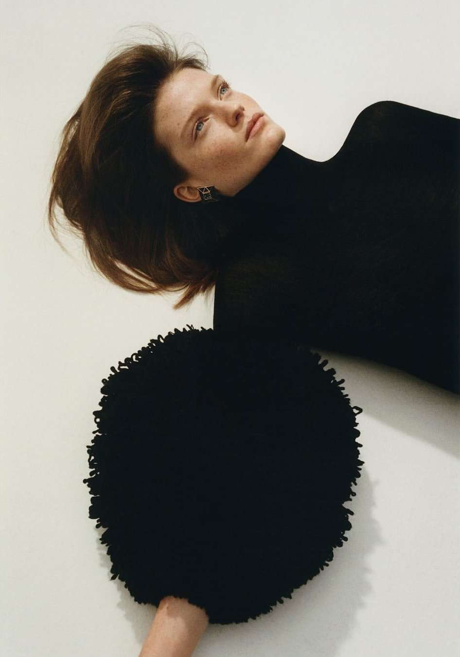 Woman in black turtleneck poses with a circular, fringed black cushion against an off-white background in a fashion editorial shot.