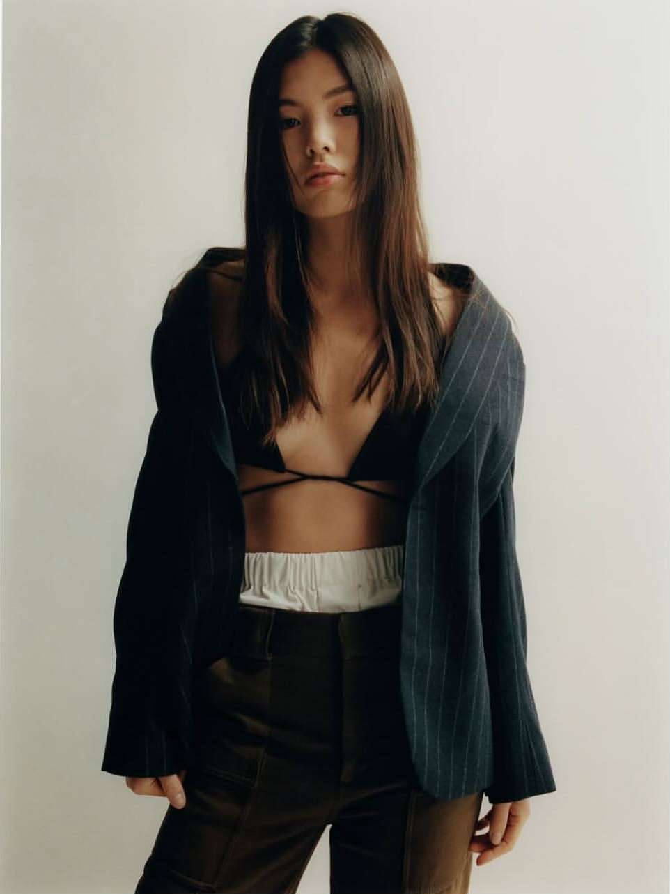 East Asian woman in an unbuttoned pinstripe blazer and dark trousers against a white backdrop.