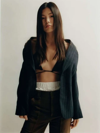 East Asian woman in an unbuttoned pinstripe blazer and dark trousers against a white backdrop.