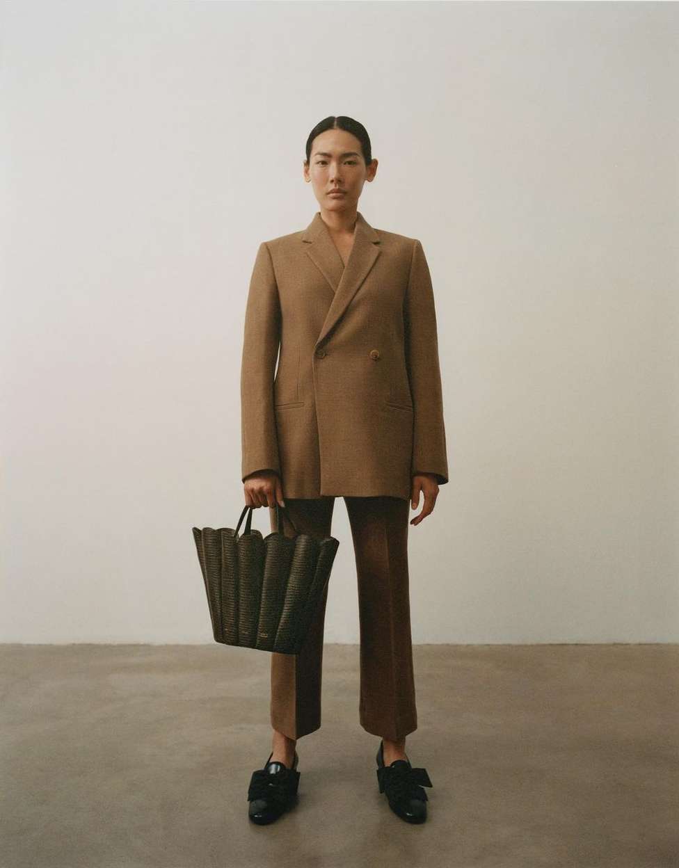 Model wearing a brown blazer and trousers holding a structured dark handbag against a plain background by Kulesza and Pik.