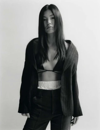 Model with long dark hair wearing an unbuttoned pinstripe blazer and dark trousers against a plain white studio backdrop.