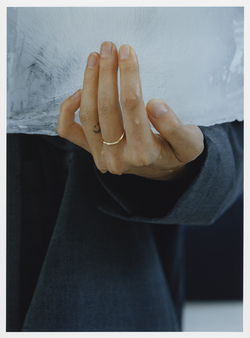 Close-up of a hand wearing a thin gold ring on the ring finger holding a block of ice with visible water droplets.
