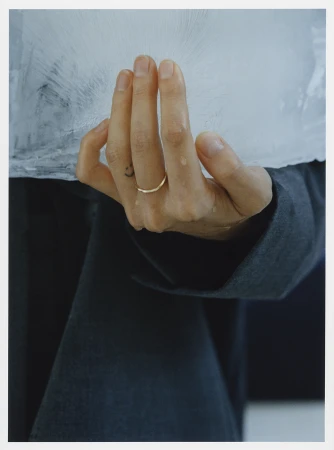 Close-up of a hand wearing a thin gold ring on the ring finger holding a block of ice with visible water droplets.