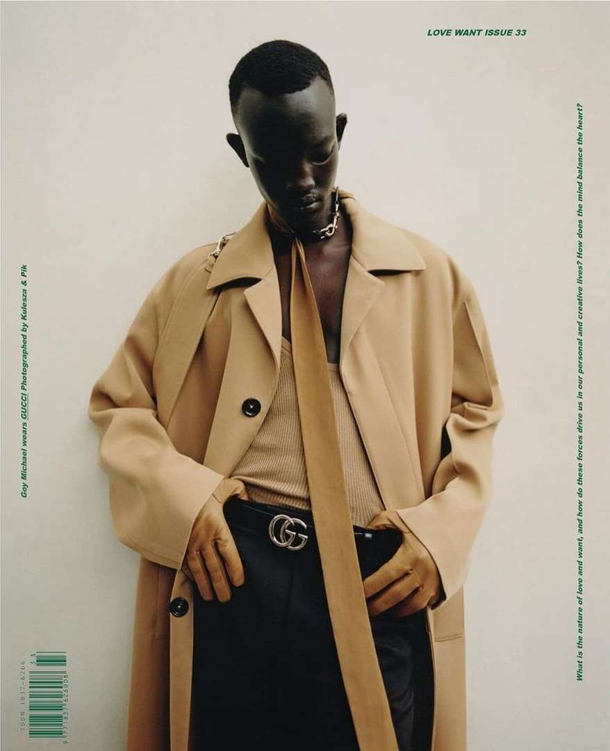 A person in a tan trench coat and black pants poses for a fashion editorial against a white wall with magazine text visible in the background.