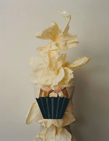 Person in artistic dress made of paper holding a dark green tote bag against a plain backdrop in a fashion or editorial style.