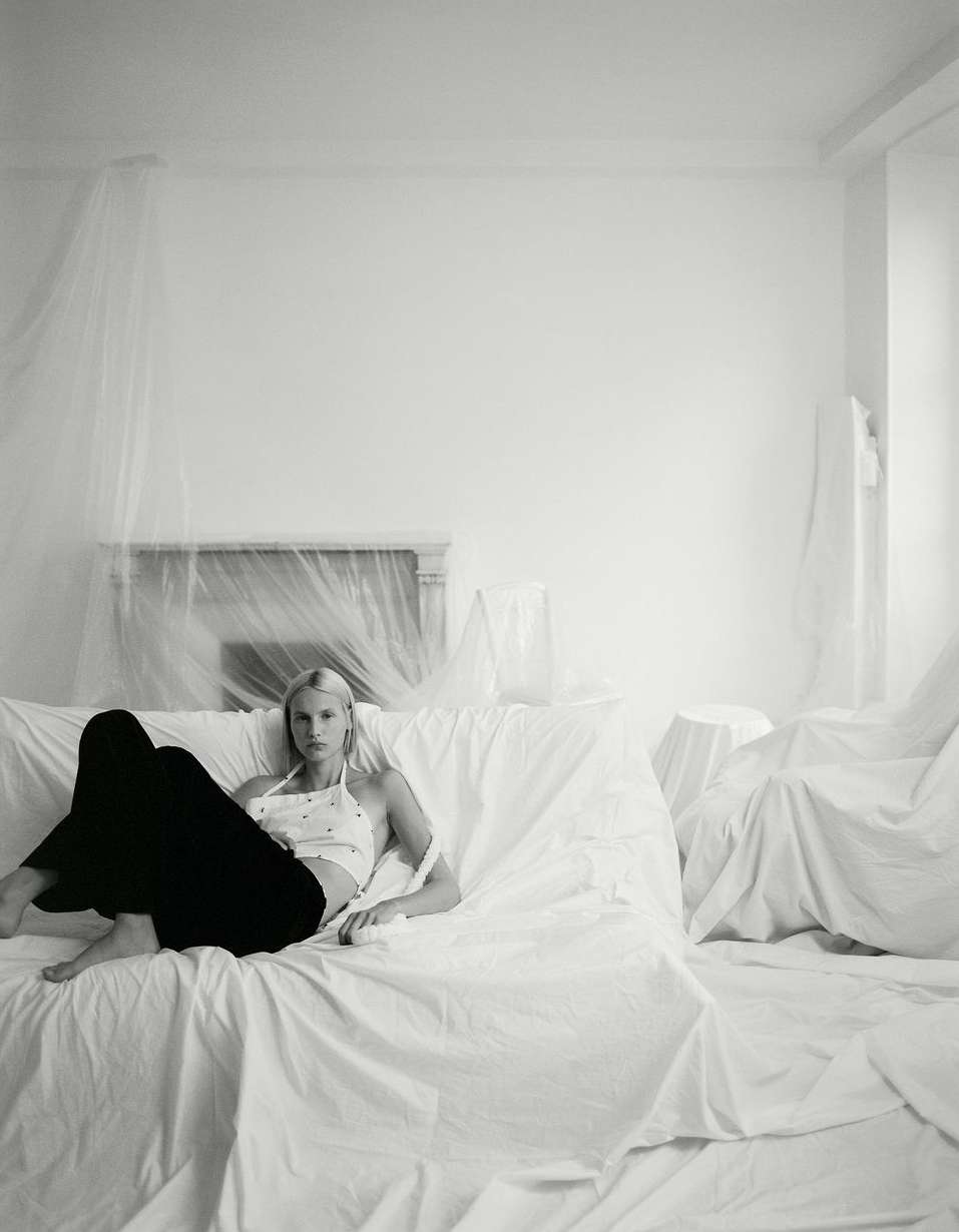 Blonde woman in halter top and dark pants reclining on a draped couch in a room with draping on the wall and fireplace.