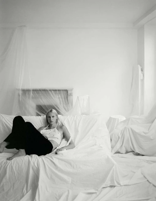 Blonde woman in halter top and dark pants reclining on a draped couch in a room with draping on the wall and fireplace.