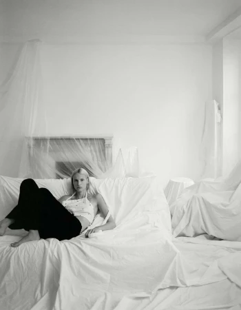 Blonde woman in halter top and dark pants reclining on a draped couch in a room with draping on the wall and fireplace.