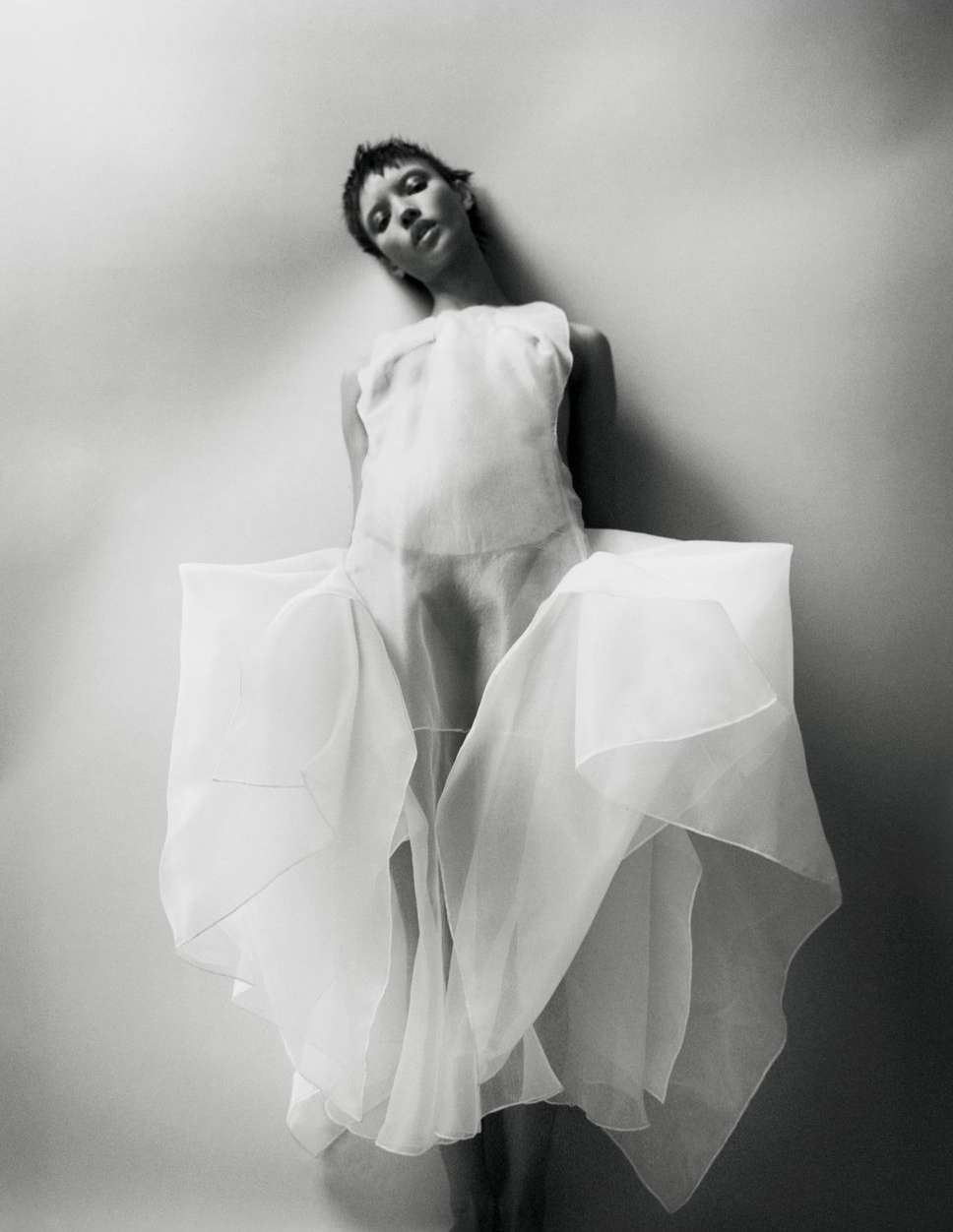 Woman in translucent white dress poses against light wall in a high-angle, black-and-white fashion portrait with soft lighting.
