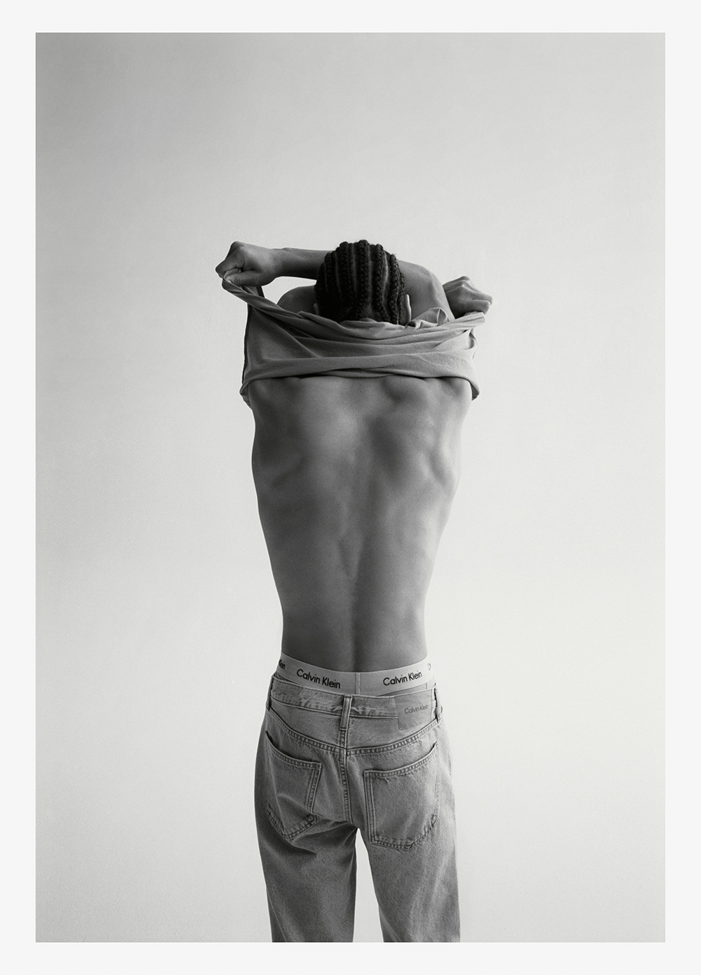 Back view of a Black adult male removing shirt; underwear band and jeans visible in a studio lit, black and white shot.