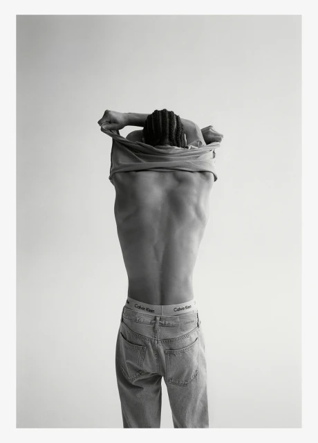 Back view of a Black adult male removing shirt; underwear band and jeans visible in a studio lit, black and white shot.