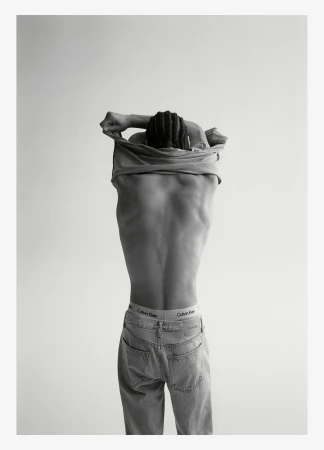 Back view of a Black adult male removing shirt; underwear band and jeans visible in a studio lit, black and white shot.