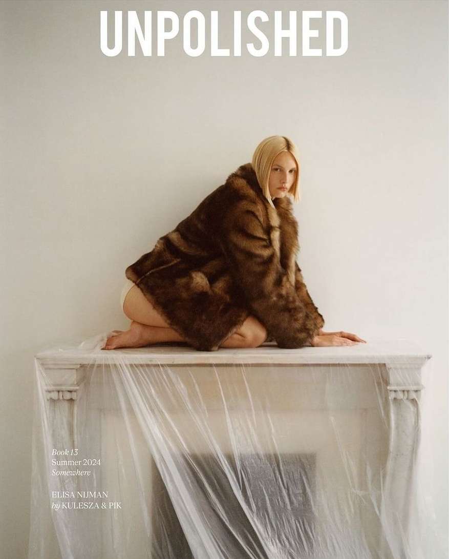 Fashion magazine cover featuring a blonde woman in a fur coat kneeling on a draped mantelpiece against a plain background.