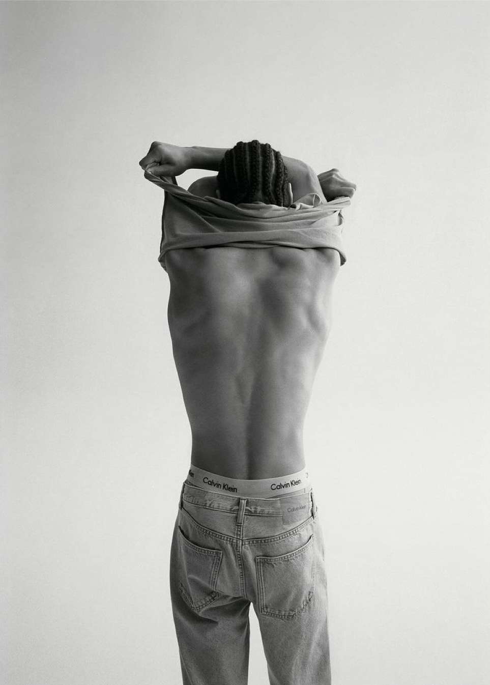 Black and white portrait of a man with an exposed back, wearing jeans, and pulling a t-shirt over his head, back to the camera.