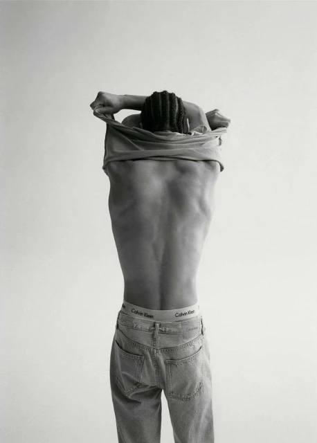 Black and white portrait of a man with an exposed back, wearing jeans, and pulling a t-shirt over his head, back to the camera.