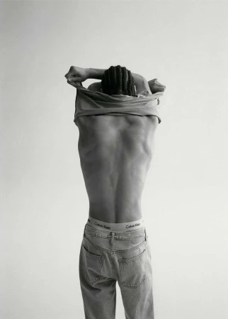 Black and white portrait of a man with an exposed back, wearing jeans, and pulling a t-shirt over his head, back to the camera.