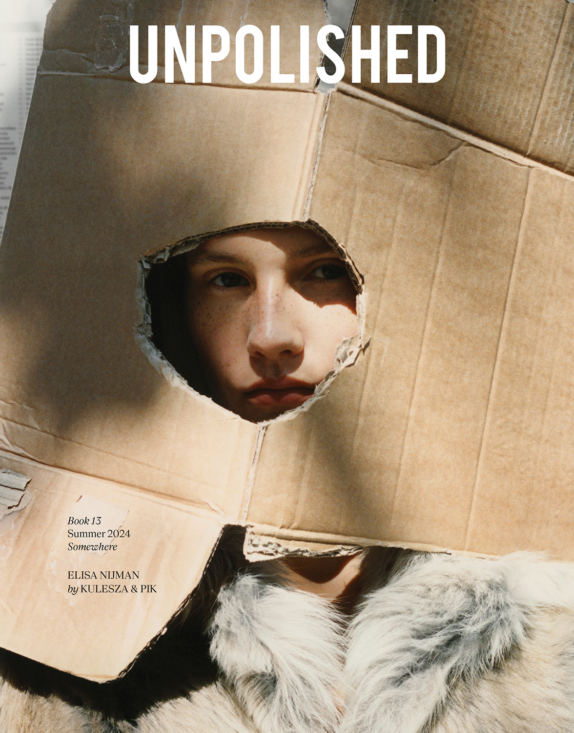 Editorial fashion shot of a fair-skinned person looking through a hole in a cardboard box, wearing a furry white coat, indoors.