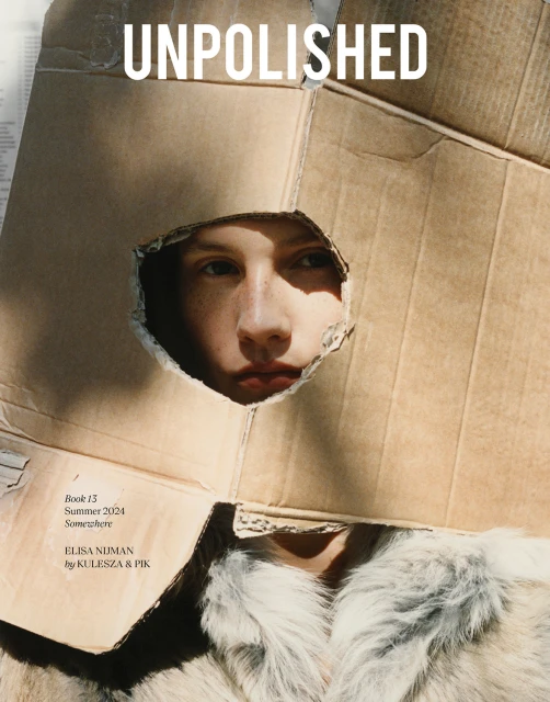 Editorial fashion shot of a fair-skinned person looking through a hole in a cardboard box, wearing a furry white coat, indoors.