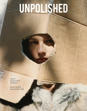 Editorial fashion shot of a fair-skinned person looking through a hole in a cardboard box, wearing a furry white coat, indoors.