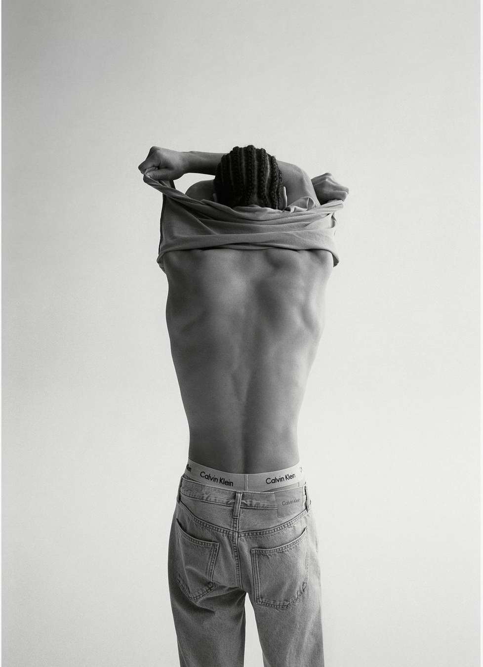 Monochrome portrait of a person taking off a t-shirt, revealing their back and jeans with a Calvin Klein waistband.