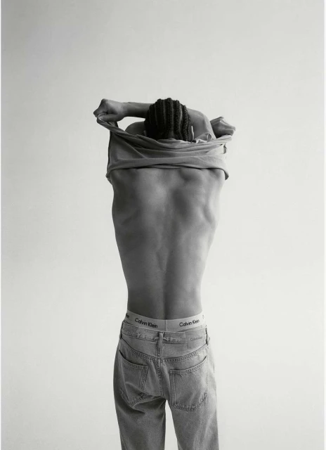 Monochrome portrait of a person taking off a t-shirt, revealing their back and jeans with a Calvin Klein waistband.