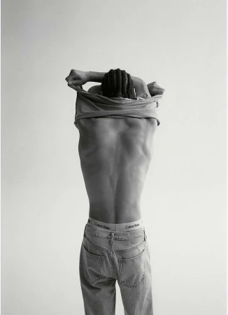 Monochrome portrait of a person taking off a t-shirt, revealing their back and jeans with a Calvin Klein waistband.