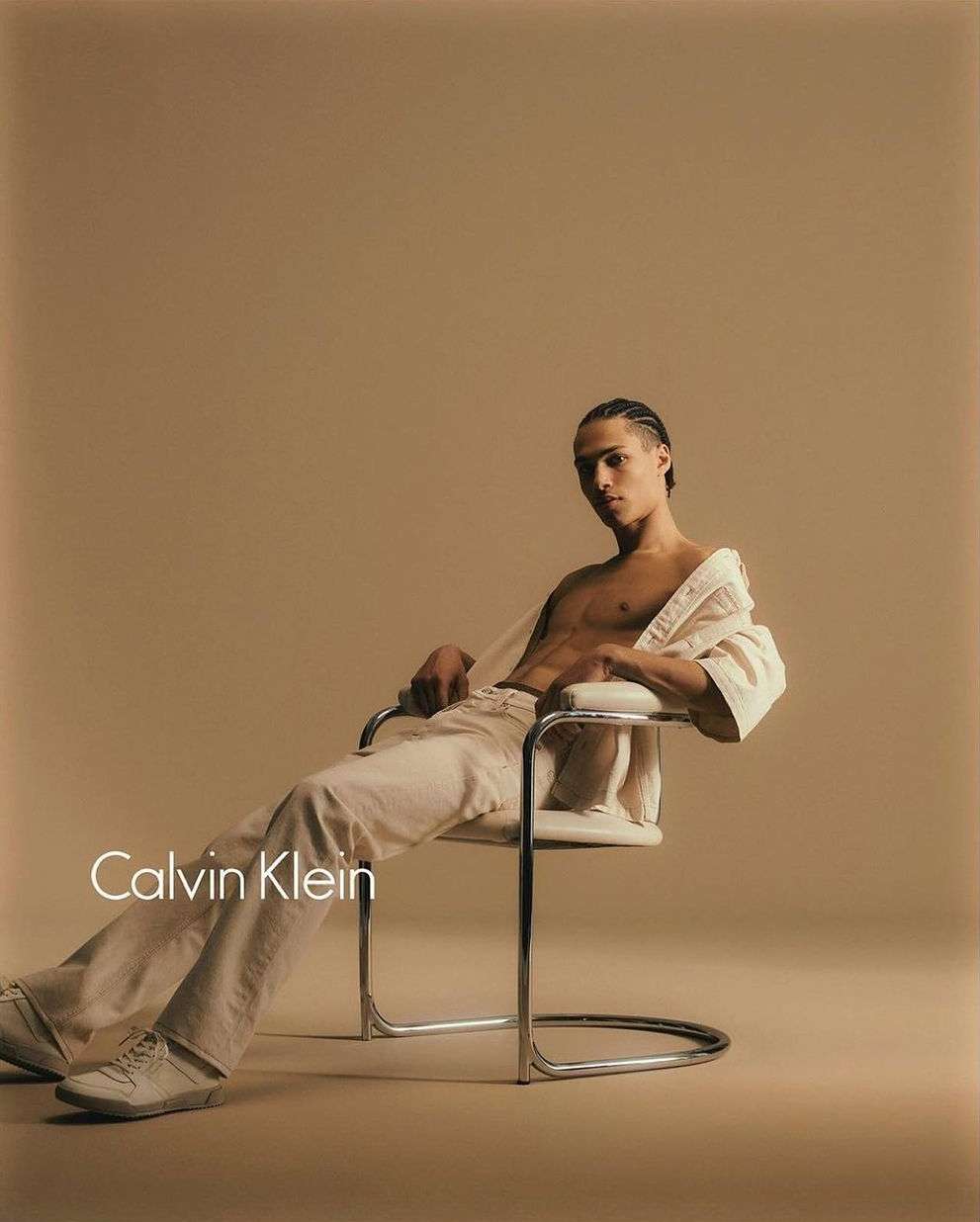 Man in white shirt and pants sits on a chair, photographed in a studio with a light brown backdrop and 'Calvin Klein' text overlay.