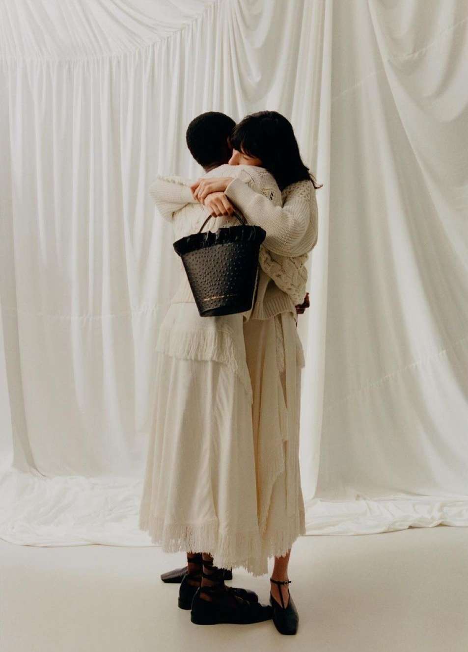 Two women embrace while modeling neutral-toned apparel and a black bucket bag in front of a textured white backdrop.