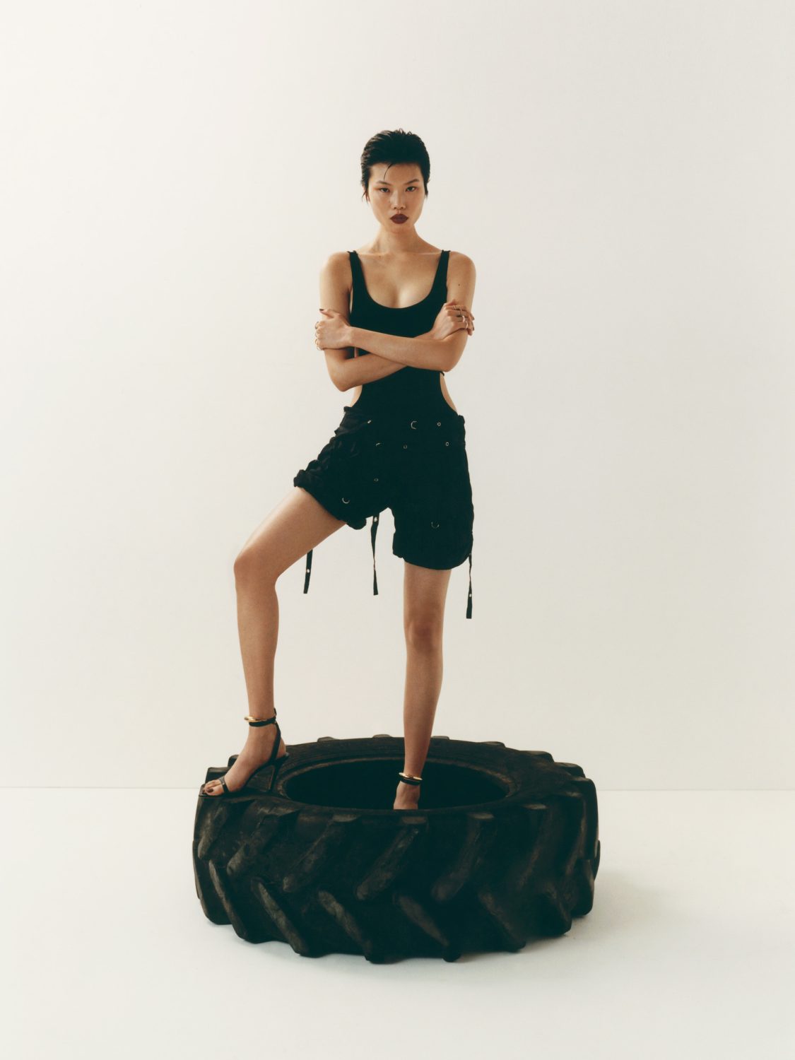 Fashion shot of a young woman in a black outfit standing in a large tire against a plain white background in a photo studio.