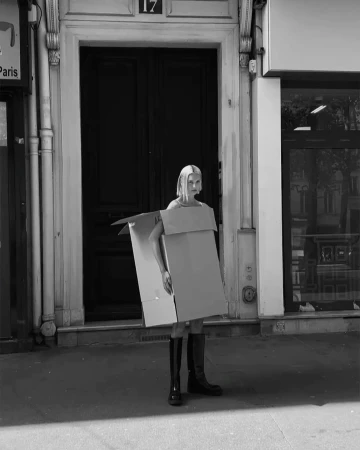 Model wearing a cardboard box costume and tall black boots standing on a city street.
