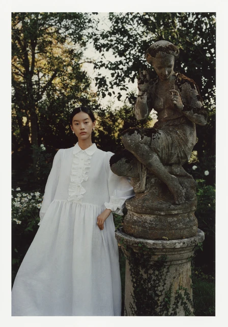 Woman in white dress poses next to weathered statue in lush garden, evoking classic elegance and natural beauty in soft daylight.