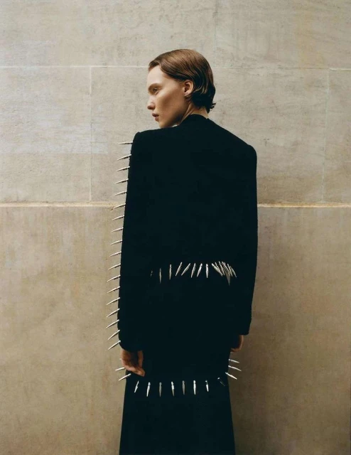 Adult posing in a black coat adorned with silver spikes, set against a muted, neutral wall; fashion-forward visual.