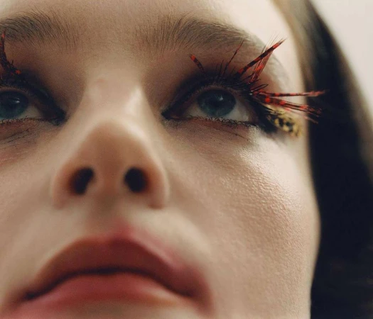 Close-up portrait of a woman looking upwards with artistic red feather eyelashes against a bright, neutral background in indoor setting.