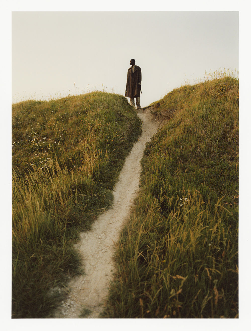 Person in a coat stands at the top of a grassy hill overlooking a path. The sky is pale, and the grass is lush in the outdoor setting.