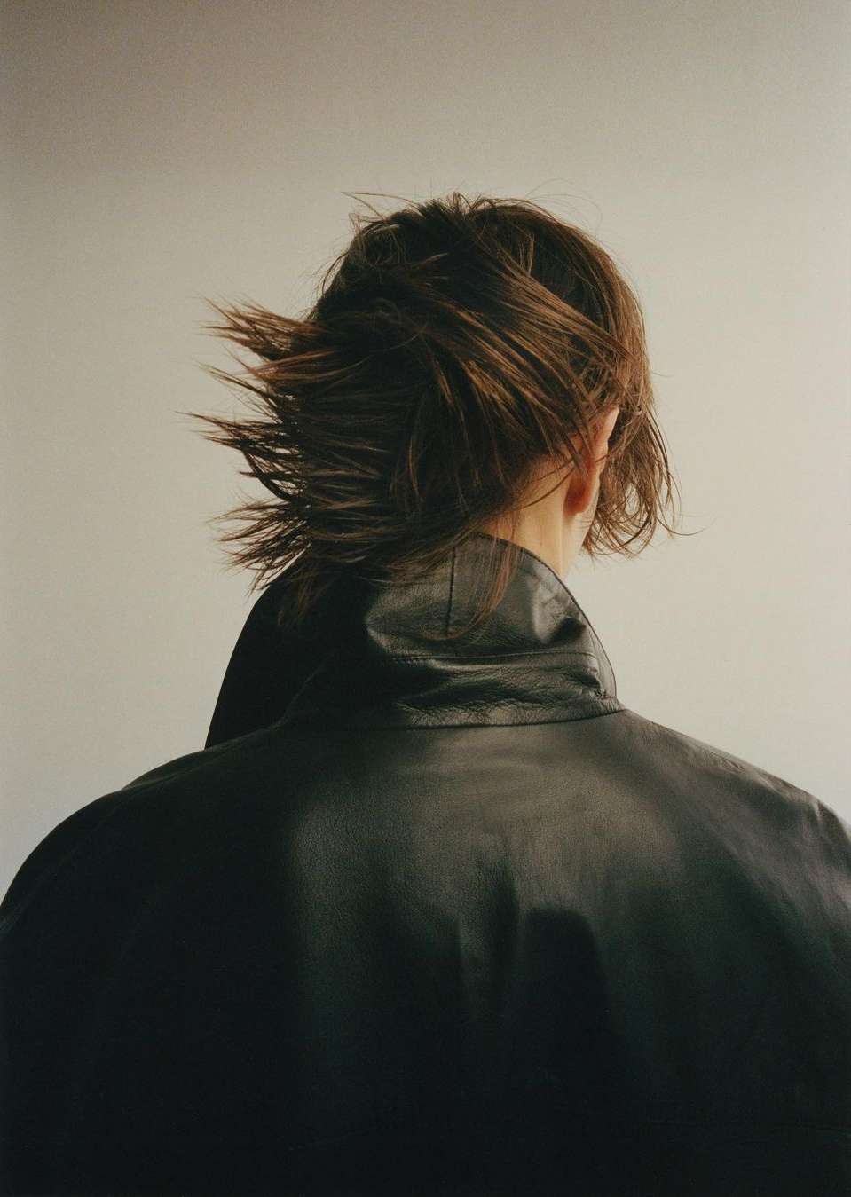 Back view of a person with short brown hair wearing a black leather jacket, captured in a studio setting with soft, diffused lighting.