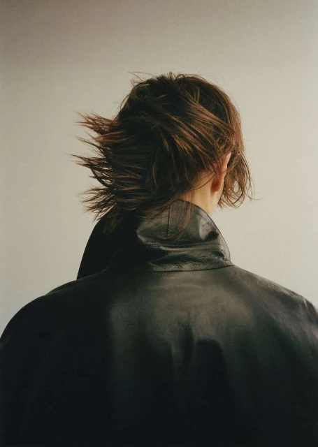 Back view of a person with short brown hair wearing a black leather jacket, captured in a studio setting with soft, diffused lighting.