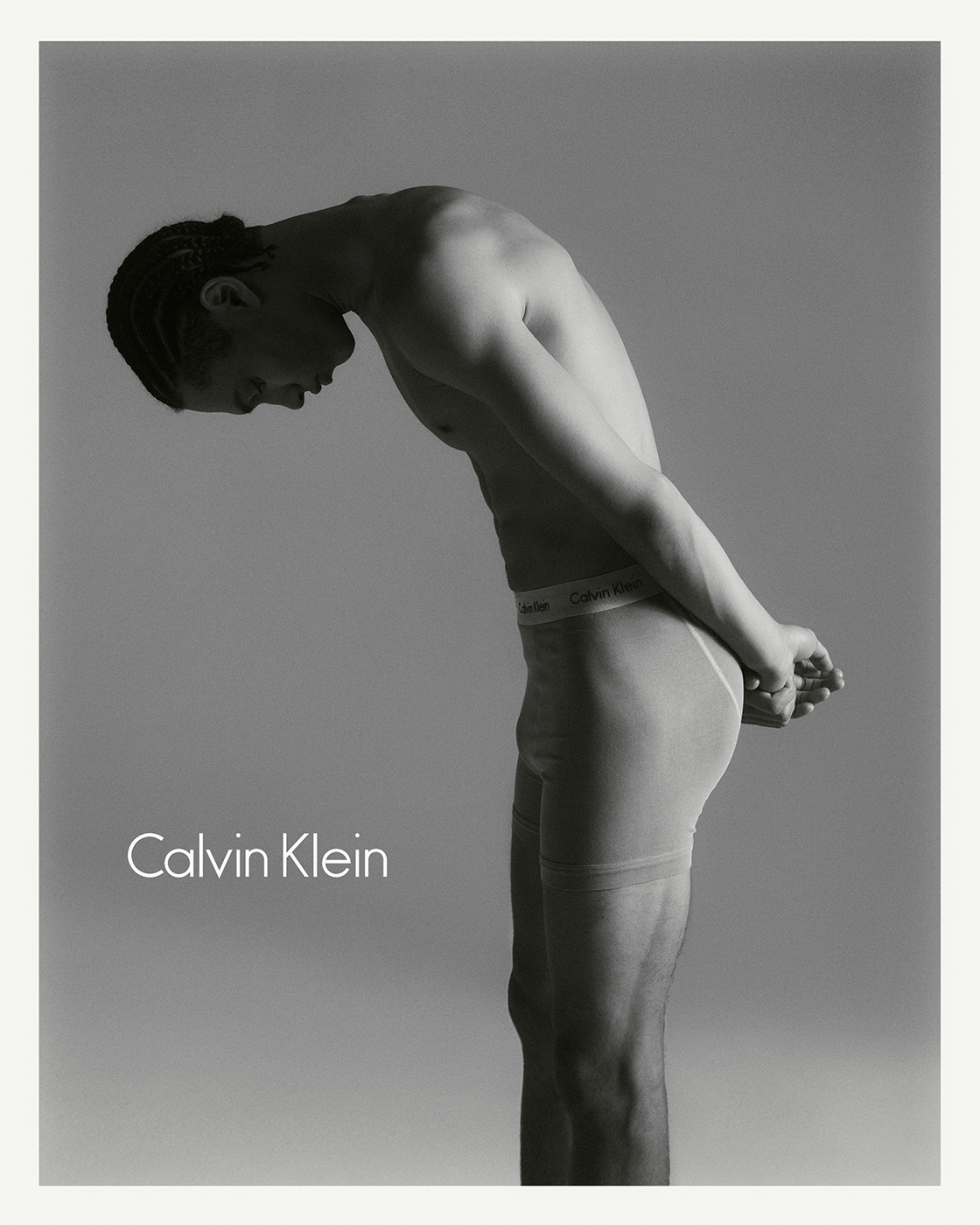 Monochrome portrait of a man in underwear bent forward against a plain grey background with Calvin Klein text