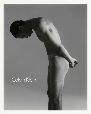 Monochrome portrait of a man in underwear bent forward against a plain grey background with Calvin Klein text