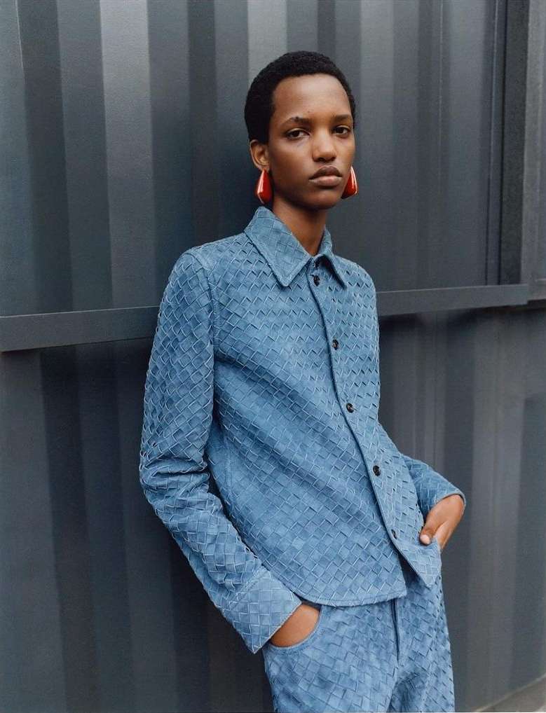 Model wearing a blue woven leather shirt and matching trousers, standing against a dark corrugated metal wall.