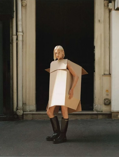 A woman in a cardboard box dress and black boots stands on a city street with stone buildings and a dark doorway in the background.