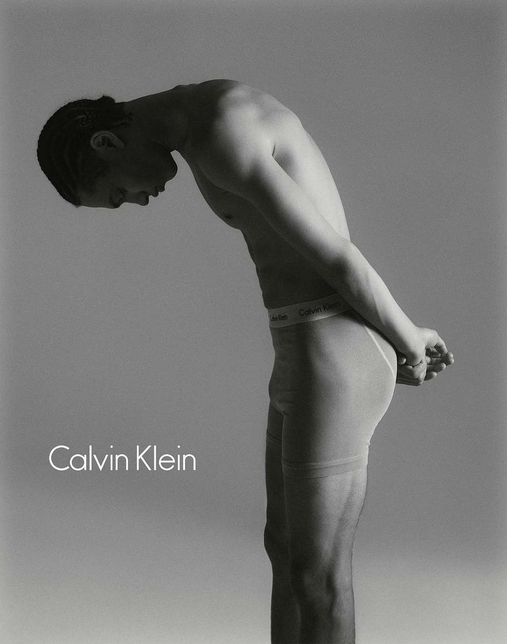 Studio shot of shirtless man in underwear, bent over with his hands behind his back, advertising Calvin Klein against grey backdrop.
