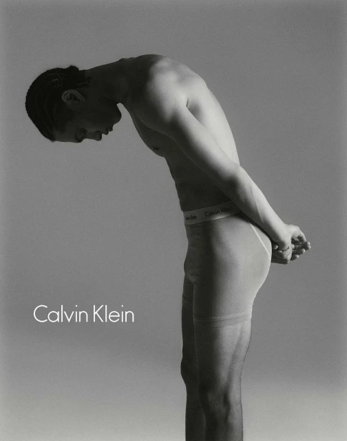 Studio shot of shirtless man in underwear, bent over with his hands behind his back, advertising Calvin Klein against grey backdrop.