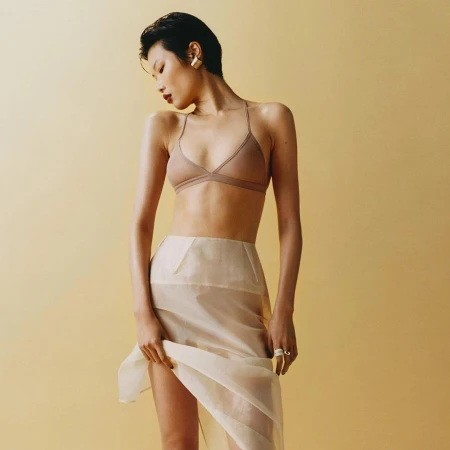 Model with short dark hair wearing a beige bralette and sheer skirt against a warm solid background