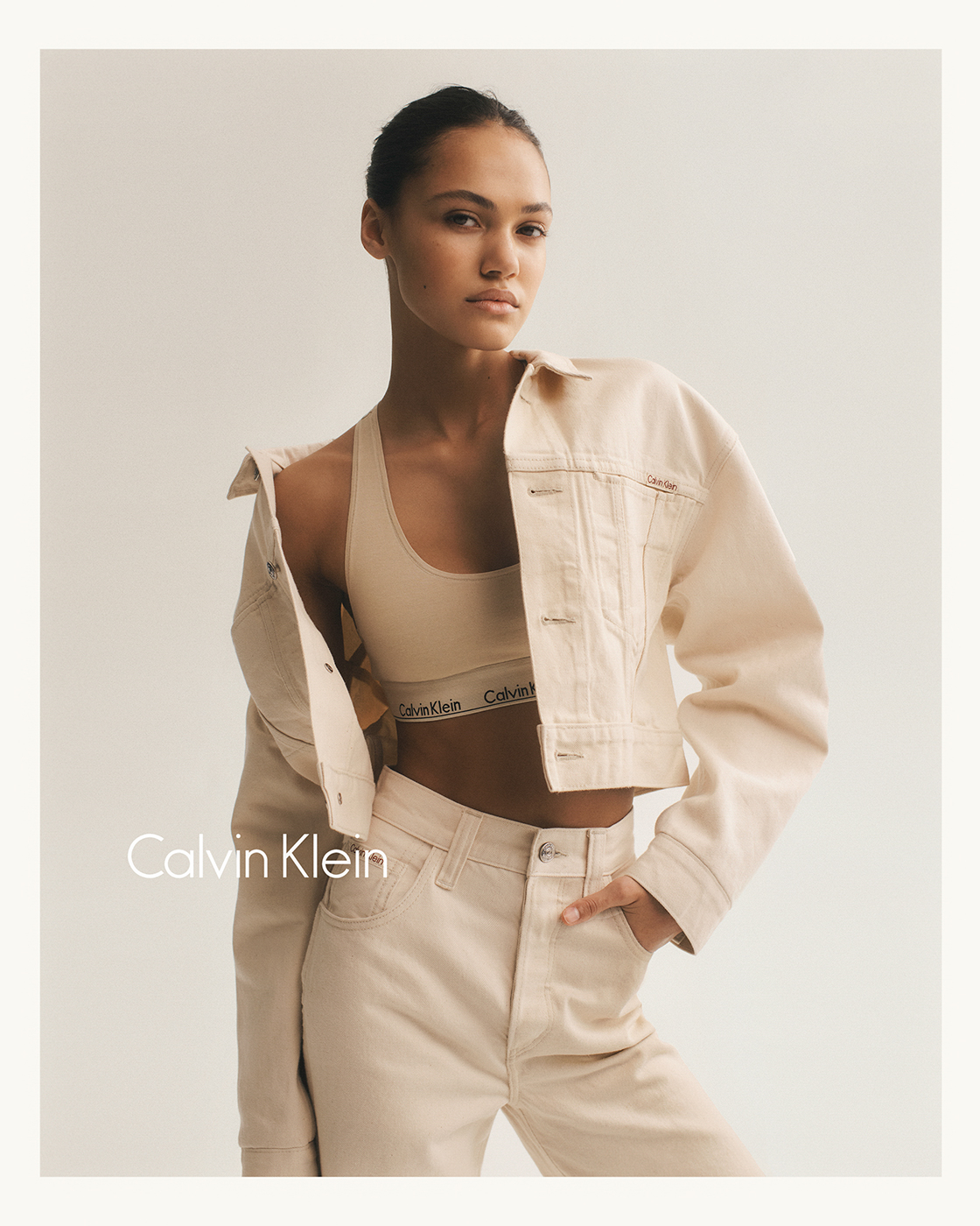 A young woman poses in Calvin Klein branded neutral athleisure and a cropped jean jacket against an off-white backdrop in a studio shot.