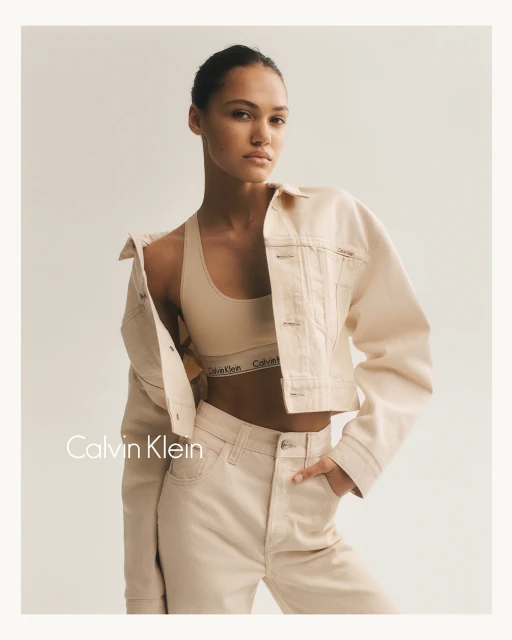 A young woman poses in Calvin Klein branded neutral athleisure and a cropped jean jacket against an off-white backdrop in a studio shot.