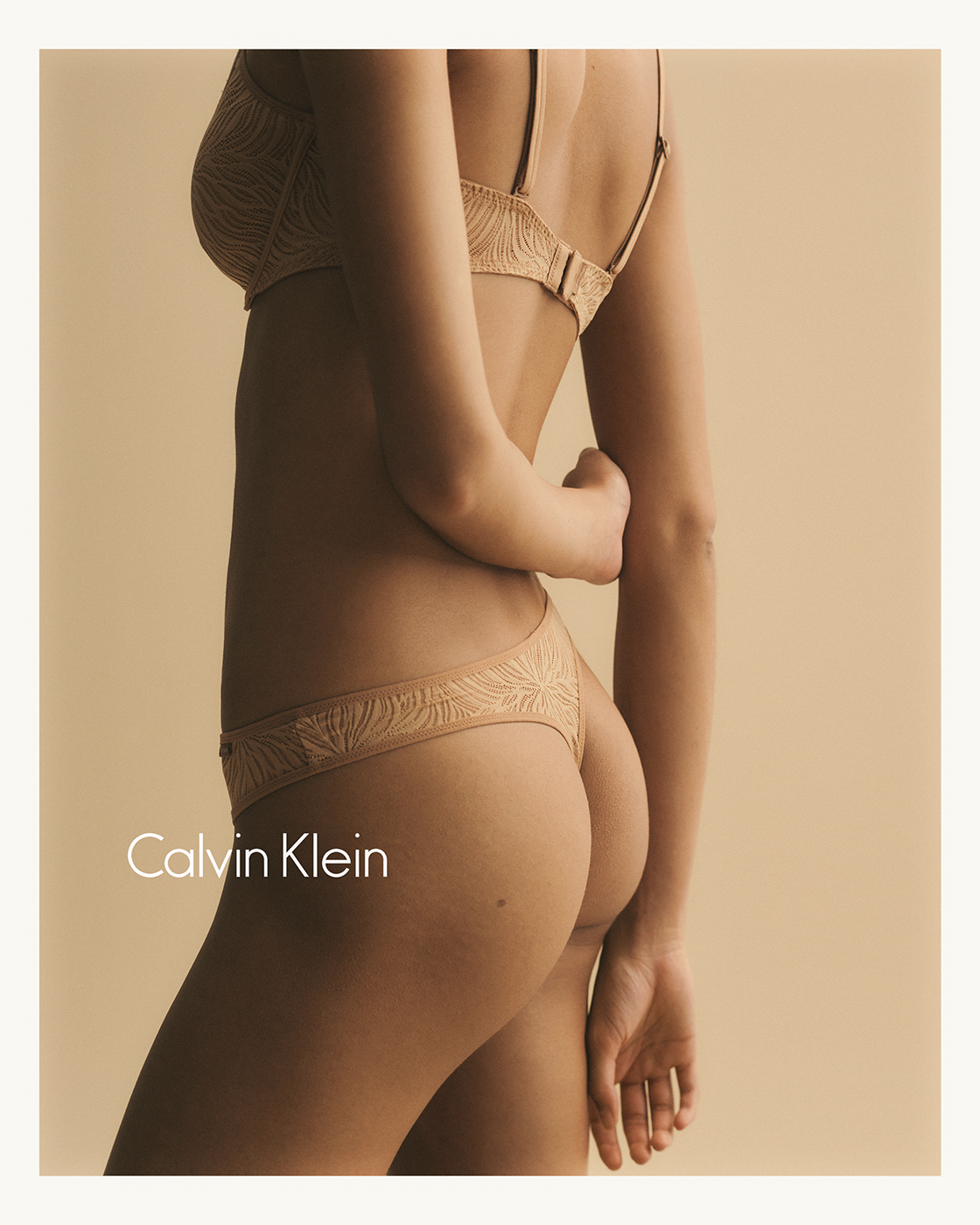 A woman in tan lingerie, standing against a neutral background, showcases the intimate apparel with Calvin Klein branding visible, medium shot.
