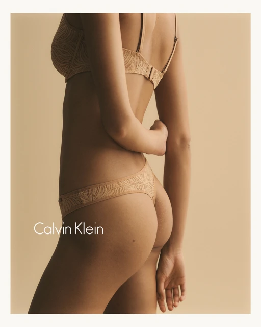 A woman in tan lingerie, standing against a neutral background, showcases the intimate apparel with Calvin Klein branding visible, medium shot.